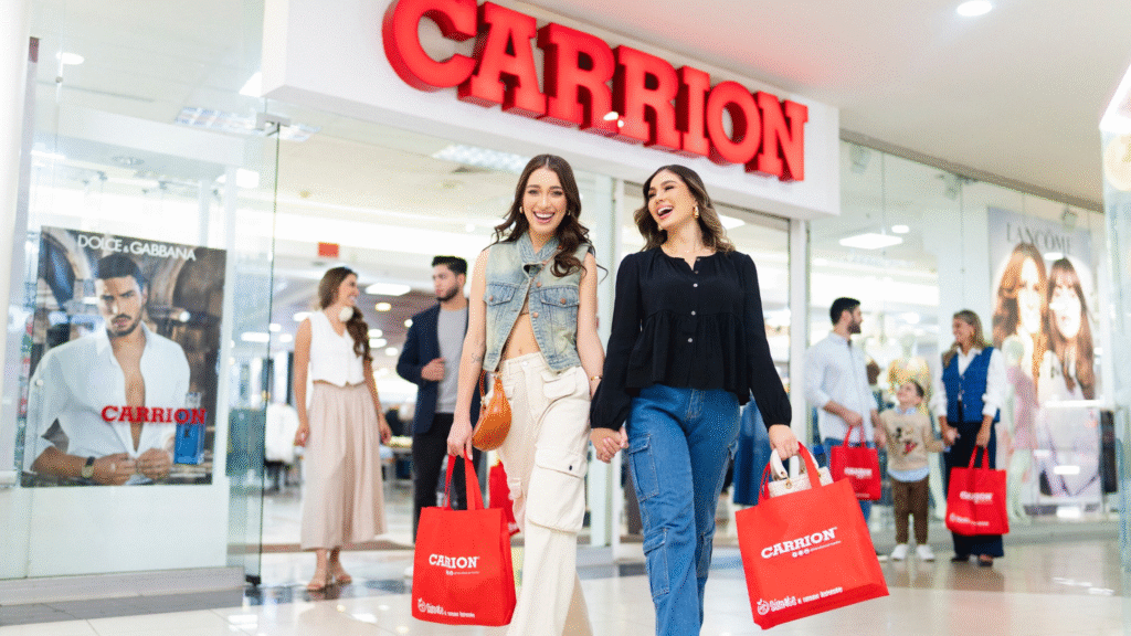 Dos mujeres jóvenes sonriendo y caminando con bolsas rojas de Tiendas Carrión en Mall Premier Catacamas.