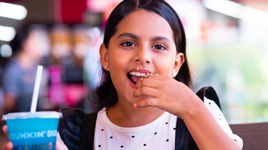 Niña sonriendo de alegría mientras come una deliciosa dona con chispas de colores de Dunkin' Donuts, uno de los postres favoritos en Mall Premier Comayagua.