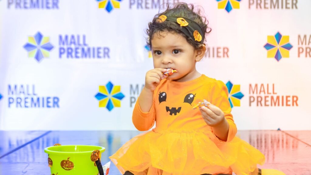Niña pequeña disfrazada con un vestido de calabaza sentada en el suelo y comiendo dulces de su balde en el evento FantasMall de Mall Premier.
