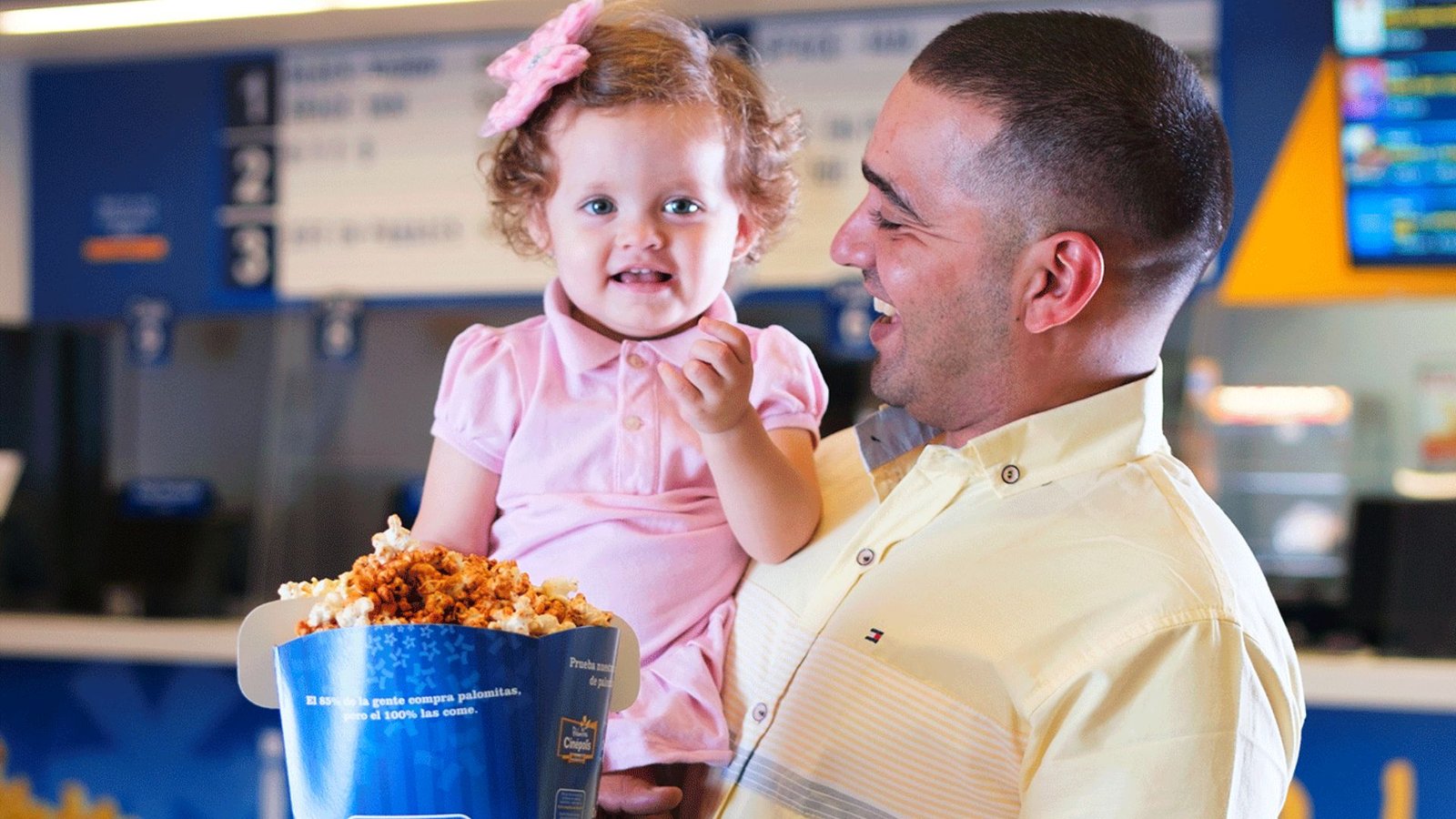 Un padre sonriente sostiene en brazos a su hija pequeña, una bebé, mientras ambos miran un balde de palomitas en el cinepolis, ubicado en Mall Premier.