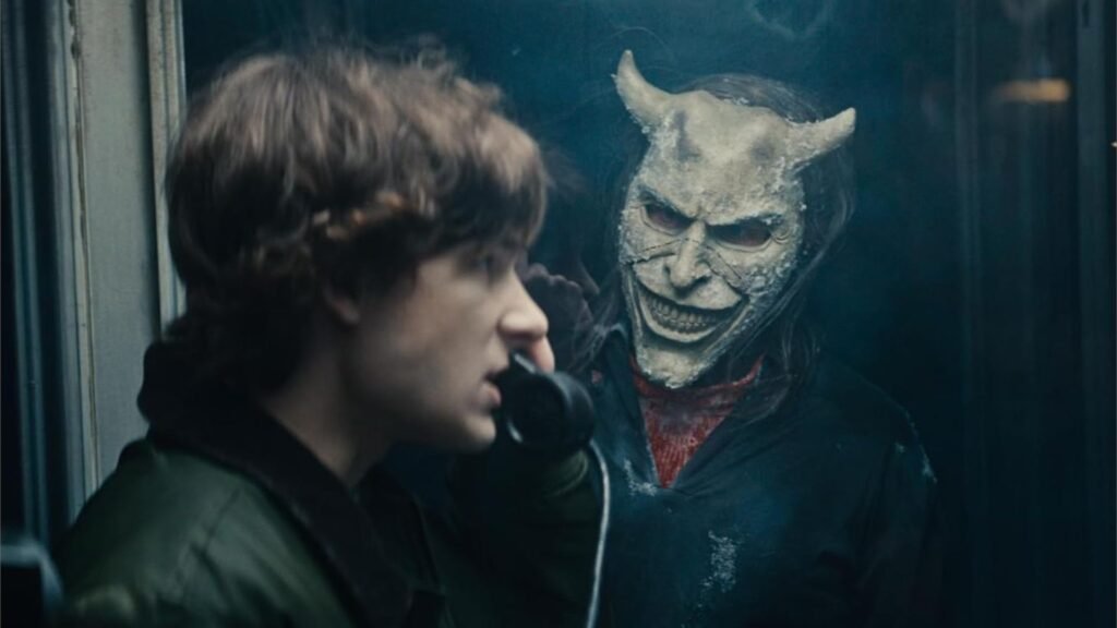 Escena de 'Teléfono Negro 2' donde un joven atiende un teléfono mientras el villano con una máscara demoníaca sonriente lo observa desde atrás.