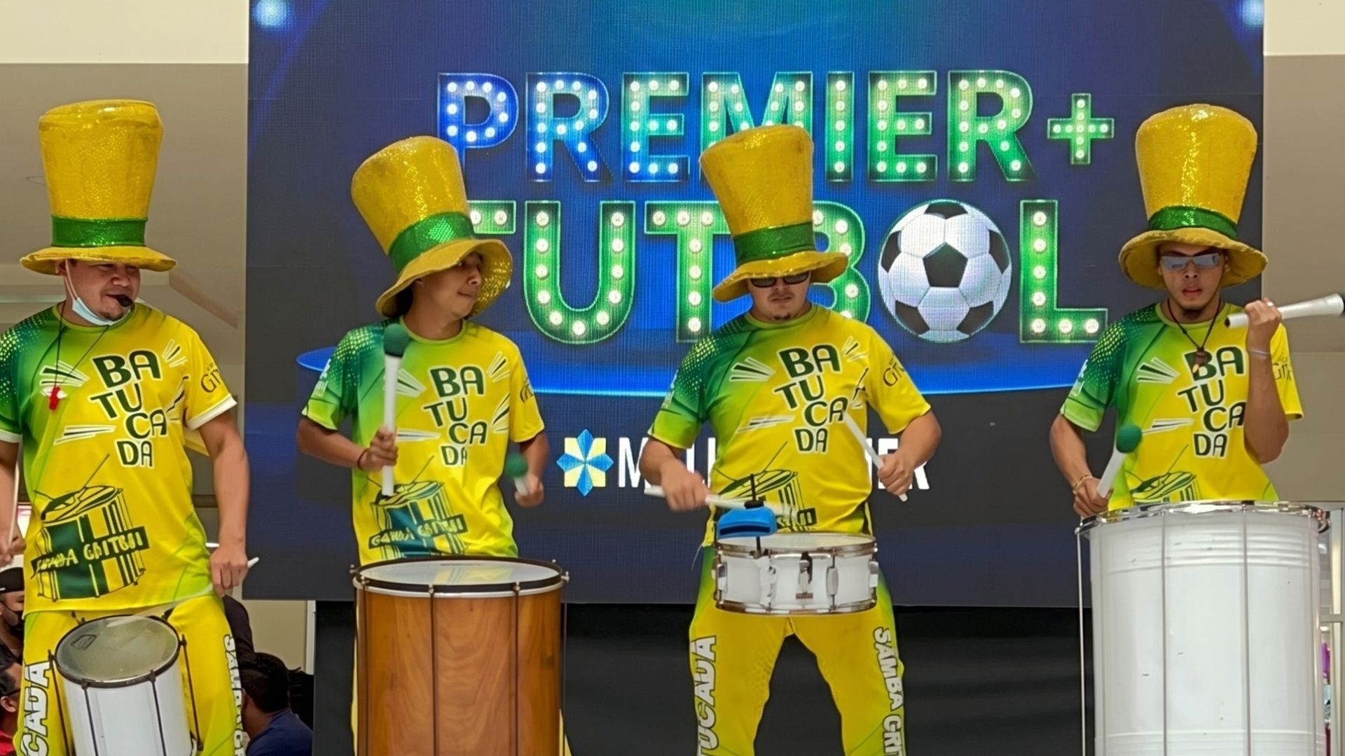 Banda de batucada con trajes amarillos y verdes y sombreros gigantes animando el Fan Fest de la inauguración del mundial en Mall Premier Tegucigalpa.