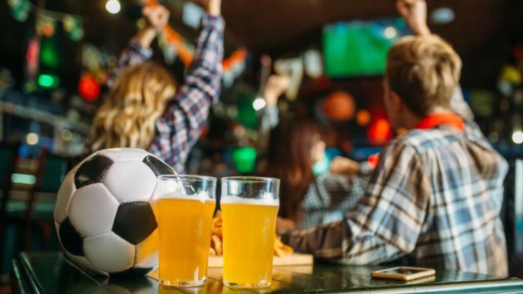 Grupo de aficionados viendo un partido del Mundial de fútbol en un bar deportivo con cervezas y un balón sobre la mesa.